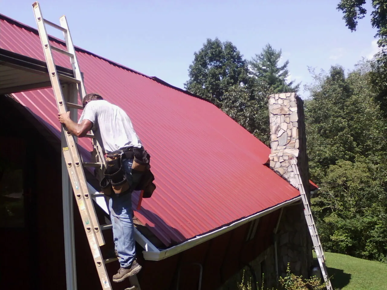 Roofer on a ladder beside a red metal roof and stone chimney — Soffit & Fascia Repair by Roofing Brothers in Sheffield Lake